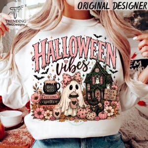 May include: A white sweatshirt featuring a graphic print with the text HALLOWEEN vibes. The design includes a ghost wearing a pink bow, a haunted house, a black cat, a stack of books labeled Basic Witch, Potions, Spells, and Magic, and pink flowers.
