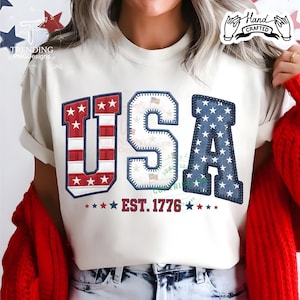 Può includere: T-shirt color crema con un design patriottico "USA". Le lettere sono rosse, bianche e blu, con stelle e strisce. Il testo "EST. 1776" è sotto. La maglietta ha un'etichetta "Hand Crafted".