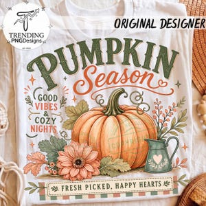 May include: A white t-shirt featuring a graphic of an orange pumpkin, a green pitcher, and a peach flower. The text reads PUMPKIN SEASON, GOOD VIBES & COZY NIGHTS, and FRESH PICKED, HAPPY HEARTS. The design includes green leaves and small berries.