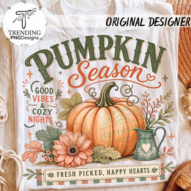 May include: A white t-shirt featuring a graphic of an orange pumpkin, a green pitcher, and a peach flower. The text reads PUMPKIN SEASON, GOOD VIBES & COZY NIGHTS, and FRESH PICKED, HAPPY HEARTS. The design includes green leaves and small berries.