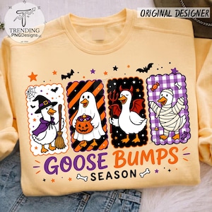 Op de afbeelding: Een lichtgeel sweatshirt met ronde hals en een afbeelding van vier eenden in Halloween-kostuums: een heks, een geest met een pompoen, een duivel en een mummie. De tekst GOOSE BUMPS SEASON staat onder de eenden, omringd door sterren en vleermuizen.