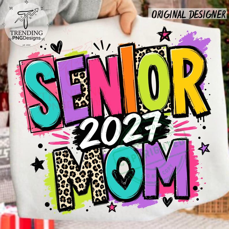 Puede incluir: Una camiseta blanca con un gr&aacute;fico colorido que muestra el texto SENIOR 2027 MOM. El dise&ntilde;o incluye letras brillantes en turquesa, morado, rosa, naranja, verde lima y amarillo, algunas con estampado de leopardo, acentuadas con estrellas negras.