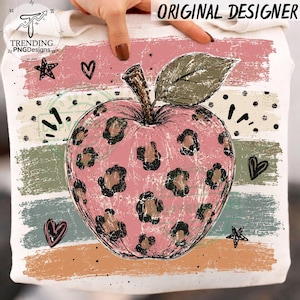 Könnte beinhalten: Ein weißes Sweatshirt mit einem großen rosa Apfel mit braunem Leopardenmuster. Das Design zeigt horizontale, abgenutzte Streifen in Rosa, Beige, Grün und Orange, akzentuiert durch schwarze Sterne, Herzen und Punkte. Der Text lautet ORIGINAL DESIGNER.