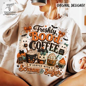 May include: A white sweatshirt featuring a graphic print with the text Freshly BOO'd COFFEE SERVED WITH Scream & Sugar. The design includes illustrations of ghosts, jack-o'-lantern coffee mugs, spiderwebs, bats, and orange polka-dot bows.