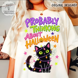 May include: A cream-colored t-shirt featuring the text "PROBABLY THINKING ABOUT HALLOWEEN" in purple, green, pink, and orange. Below the text is a black cat wrapped in colorful string lights, surrounded by small pink, yellow, and orange stars.