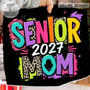 Puede incluir: Un dise&ntilde;o gr&aacute;fico negro con el texto SENIOR 2027 MOM en letras llamativas y coloridas. Las letras incorporan colores rosa brillante, turquesa, amarillo y verde lima con patrones de leopardo, salpicaduras de pintura y estrellas.