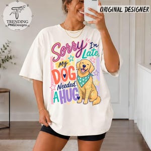 May include: An off-white t-shirt featuring a graphic print of a golden retriever wearing a teal bandana with white hearts. The text reads Sorry I'm Late, My DOG Needed A HUG, surrounded by colorful stars and vertical stripes in pink, yellow, and blue.