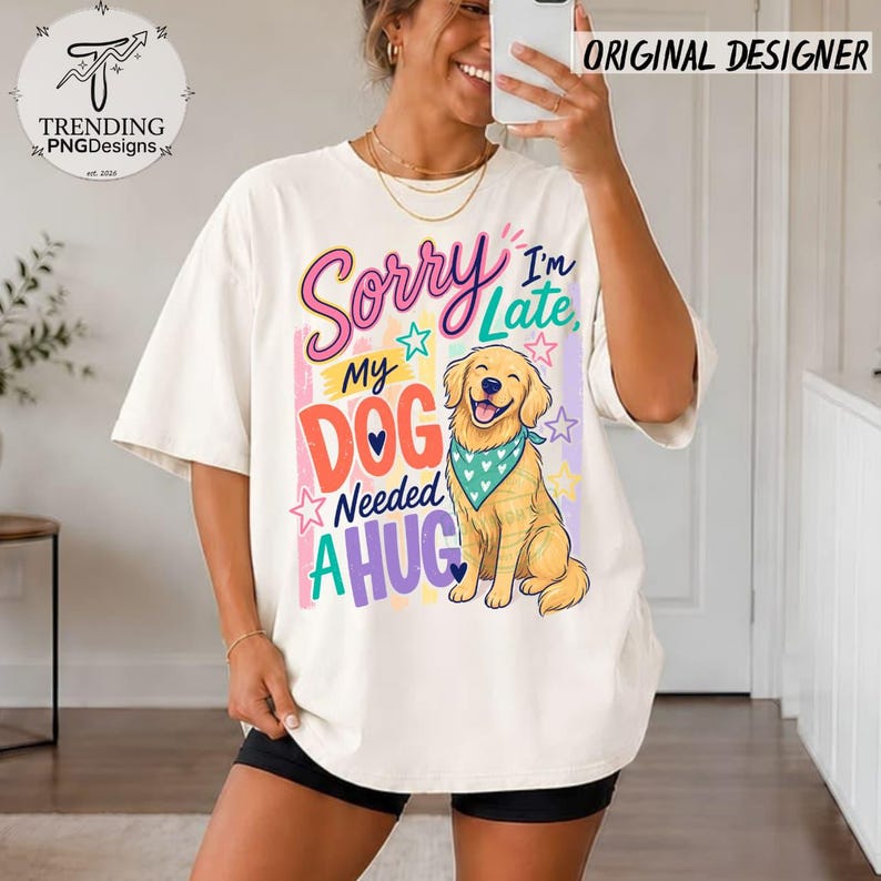 May include: An off-white t-shirt featuring a graphic print of a golden retriever wearing a teal bandana with white hearts. The text reads Sorry I'm Late, My DOG Needed A HUG, surrounded by colorful stars and vertical stripes in pink, yellow, and blue.