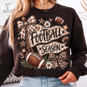 May include: A dark brown sweatshirt featuring a graphic print with the text "FOOTBALL SEASON" in a brown script font. The design includes a brown and white plaid bow, a leopard print bow, footballs, white daisies, and various star shapes.