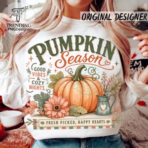 May include: A white sweatshirt featuring a graphic of an orange pumpkin, a peach flower, and a green pitcher. Text reads: PUMPKIN SEASON, GOOD VIBES & COZY NIGHTS, and FRESH PICKED, HAPPY HEARTS. The design includes green leaves and small berries.