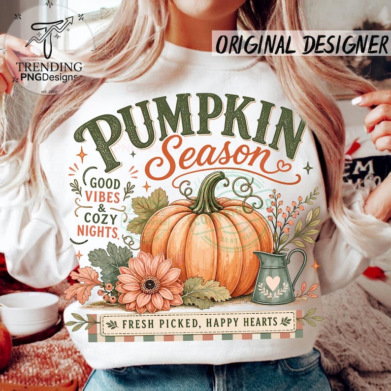 May include: A white sweatshirt featuring a graphic of an orange pumpkin, a peach flower, and a green pitcher. Text reads: PUMPKIN SEASON, GOOD VIBES & COZY NIGHTS, and FRESH PICKED, HAPPY HEARTS. The design includes green leaves and small berries.