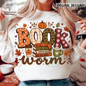 May include: A white sweatshirt featuring a graphic print with the text BOOK worm. The design includes a pumpkin, a stack of three books labelled COZY DAYS, PUMPKIN SPICE, and FALL VIBES, a coffee cup, a mug with whipped cream, and autumn leaves.