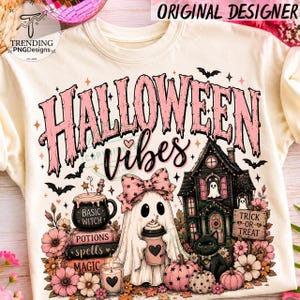 May include: A cream-colored t-shirt featuring a graphic print with the text HALLOWEEN VIBES in pink. The design includes a ghost wearing a pink bow, a black cat, a haunted house, a stack of books labeled POTIONS SPELLS MAGIC, and flowers.