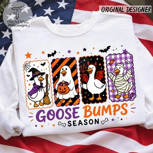 Op de afbeelding: Een wit sweatshirt met lange mouwen en een afbeelding van vier ganzen in Halloween-kostuums: een heks, een geest met een pompoen, een duivel en een mummie. De tekst "GOOSE BUMPS SEASON" staat in paars en oranje onder de ganzen gedrukt.