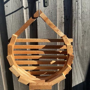 May include: A wooden hanging planter with a geometric design. The planter is constructed from light brown wood and features a slatted basket-like structure. It is suspended by a metal wire and mounted on a wooden fence.