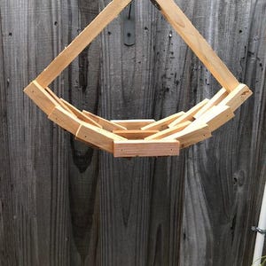 May include: A hanging planter constructed from light-colored natural wood slats arranged in a geometric, tiered, semi-circular basket shape. The structure is suspended by a metal hook and wire, featuring a series of interlocking wooden pieces.