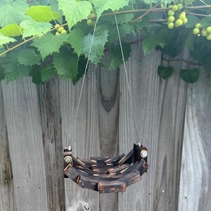 May include: A hanging bird feeder constructed from charred, dark-stained wooden blocks arranged in a curved, basket-like shape. The structure is suspended by two thin metal wires attached to the sides with silver-colored hardware bolts.