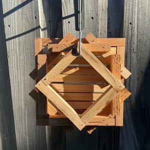 May include: A square, wooden hanging planter with a geometric design. The planter is constructed from light brown wood and features a diamond-shaped frame within the square. It is suspended by a metal hanger, perfect for displaying plants.
