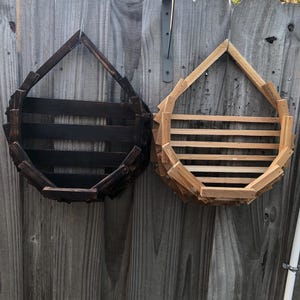 Könnte beinhalten: Zwei hängende Holzpflanzgefäße, eines dunkelbraun gebeizt und das andere aus Naturholz, sind an einem verwitterten grauen Holzzaun befestigt. Jeder Pflanztopf hat eine tropfenförmige Form mit horizontalen Lamellen.