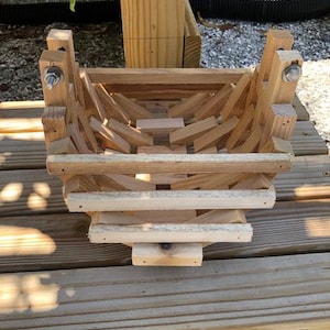 May include: A square-shaped basket constructed from light-colored wooden slats arranged in a crisscross lattice pattern. The structure features vertical corner posts secured with metal bolts and nuts at the top, creating an open-frame design.