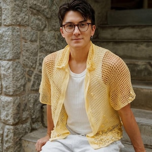 May include: A pale yellow short-sleeved shirt featuring an open-knit crochet texture with a collared neckline and button-down front, worn over a plain white ribbed tank top paired with light-colored trousers and dark-rimmed eyeglasses.