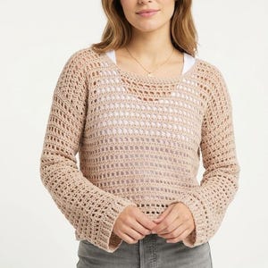 May include: A light beige, open-weave crochet sweater with long sleeves and a scoop neckline. The sweater is worn over a white top and paired with gray pants. The crochet design creates a textured, airy look. The sweater is a relaxed fit.
