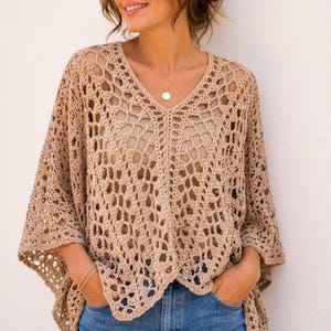 Pode incluir: Um poncho de crochê bege com um padrão de losangos vazados, decote em V e mangas largas. A peça é usada sobre uma blusa bege lisa e calças jeans azuis. O design de crochê cria uma aparência texturizada e arejada com bordas festonadas.