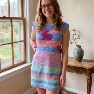 May include: A colorful, handmade crochet dress with horizontal stripes in pastel shades of blue, pink, purple, and green. The top features a blue background with a pink bunny design. The dress has tie details on the sides.
