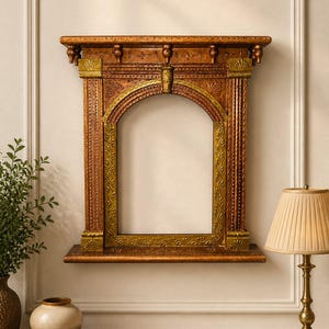 Op de afbeelding: Een sierlijke, houten wandframe met een warme bruine afwerking en gouden accenten. Het frame heeft een boogvormige opening en decoratieve gravures, wat een vintage uitstraling creëert. Het is aan een witte muur bevestigd. Een lamp en andere items staan op de voorgrond.