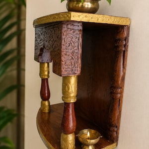 K&ouml;nnte beinhalten: Ein wandmontiertes Holzregal aus dunkelbraunem Holz mit goldfarbenen Akzenten, aufwendigen Blumenverzierungen am oberen Rahmen und zwei gedrechselten S&auml;ulen. Oben steht eine kleine Messingschale mit einer gr&uuml;nen Pflanze, unten eine brennende &Ouml;llampe.