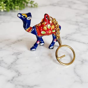 Op de afbeelding: Een kameelvormige sleutelhanger met een felblauw lichaam en handgeschilderde details in rood, goud en wit. De kameel heeft een decoratief zadelontwerp op de rug en is bevestigd aan een goudkleurige metalen ketting met een ronde sleutelring.