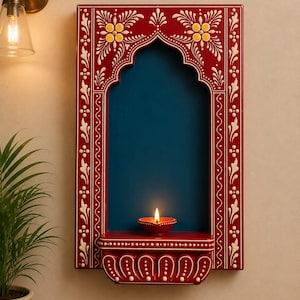 Puede incluir: Un estante rectangular de pared con un marco rojo intenso con patrones florales y geométricos blancos que rodean un nicho arqueado azul oscuro. Una pequeña lámpara de aceite de arcilla roja con una llama encendida descansa sobre el estante.