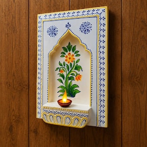 Peut inclure: Une étagère murale rectangulaire présentant une niche arquée blanche avec un motif floral peint à la main en orange et vert. Le cadre est orné de motifs bleus et jaunes. Une petite lampe diya en terre cuite brune avec une flamme allumée est posée.