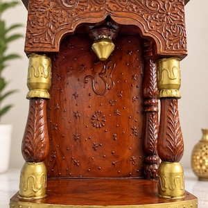 K&ouml;nnte beinhalten: Ein h&ouml;lzerner Heimtempel mit einem satten braunen Finish, aufwendigen Blumen-Schnitzereien und goldfarbenen Akzenten. Die Struktur umfasst zwei verzierte S&auml;ulen, ein zentrales Om-Symbol und eine kleine h&auml;ngende Glocke auf einem stabilen Sockel.