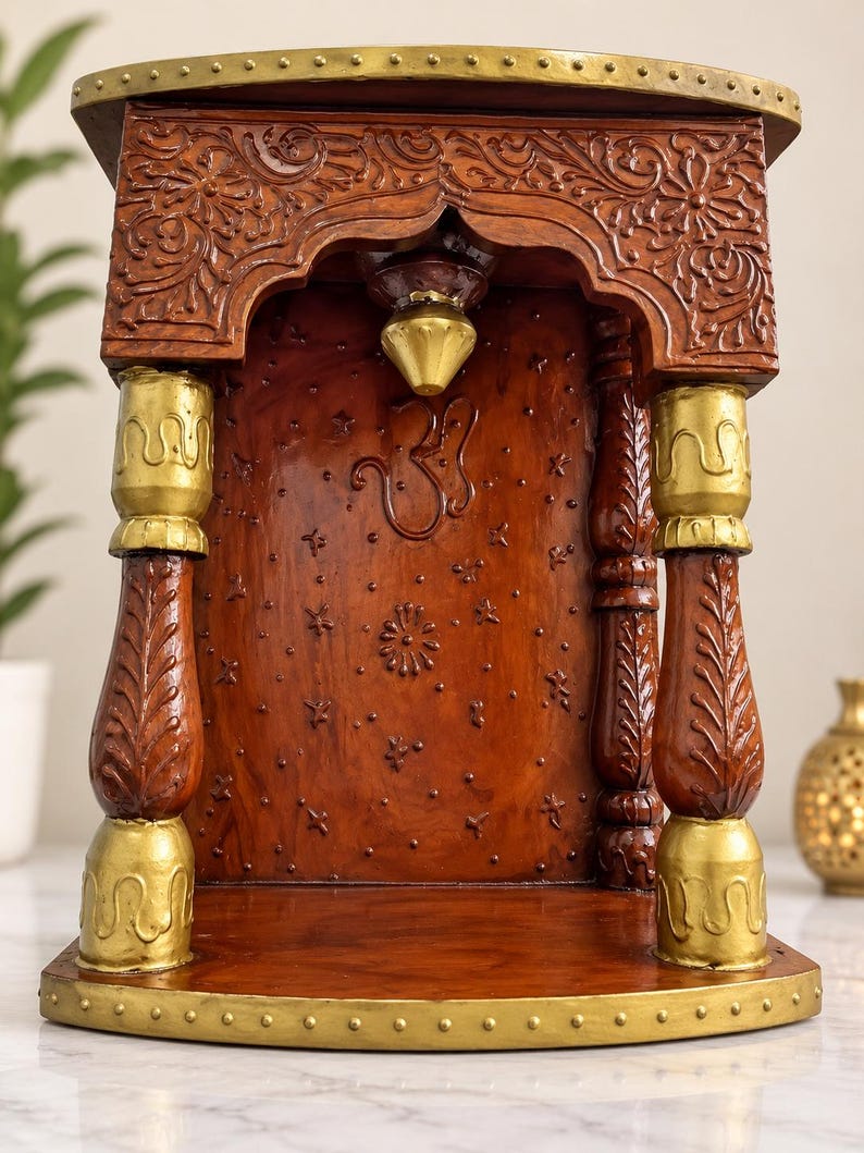 K&ouml;nnte beinhalten: Ein h&ouml;lzerner Heimtempel mit einem satten braunen Finish, aufwendigen Blumen-Schnitzereien und goldfarbenen Akzenten. Die Struktur umfasst zwei verzierte S&auml;ulen, ein zentrales Om-Symbol und eine kleine h&auml;ngende Glocke auf einem stabilen Sockel.