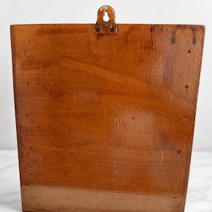 K&ouml;nnte beinhalten: Ein rechteckiger Wandhalter aus Holz mit polierter, mittelbrauner Oberfl&auml;che. Das St&uuml;ck weist eine sichtbare Holzmaserung, eine flache Vorderseite und eine Metallaufh&auml;ngung auf, die mit zwei kleinen Schrauben oben mittig befestigt ist.