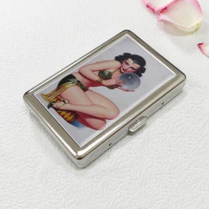 May include: A silver metal cigarette case featuring a vintage pin-up illustration. The artwork depicts a woman in a green and gold outfit holding a crystal ball. The case has a rectangular shape with a hinged lid and a clasp closure.