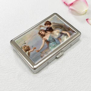 May include: A silver metal cigarette case featuring a classical painting of three women and a cherub. The artwork depicts figures in flowing gowns with a soft, pastel color palette. The case has a rectangular shape with a hinged closure.