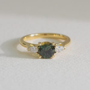 May include: A gold ring featuring a round, dark green center stone held by four prongs. The center stone is flanked on each side by a single, smaller, clear, pear-shaped stone. The band is smooth, polished yellow gold with a simple, classic design.