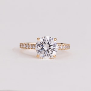 May include: A gold ring featuring a large, round, brilliant-cut clear center stone held by four prongs. The band is crafted from yellow gold and is accented with a row of small, round, clear pavé-set stones along both sides of the shank.