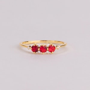 May include: A gold ring featuring three round, vibrant red stones set in a horizontal row, accented by four small, clear, round stones placed between and at the ends of the red stones, all held in place by a thin, polished gold band.
