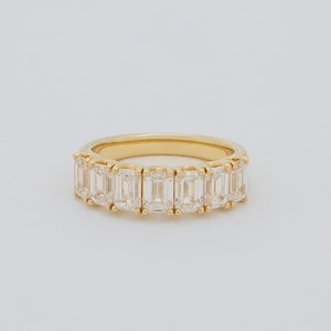 May include: A gold band ring featuring a row of seven clear emerald-cut stones set in prongs. The ring has a polished yellow gold finish and a continuous design where the stones are arranged side by side along the front half of the band.