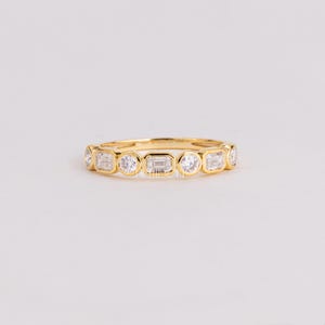 May include: A gold band ring featuring an alternating pattern of bezel-set round and emerald-cut clear stones. The metal has a polished yellow finish, and the stones are arranged in a continuous row along the top half of the circular band.