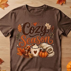 May include: A brown short-sleeve t-shirt featuring a graphic with the text Cozy Season in dark brown and orange script. The design includes a white mug with a heart, a small orange pumpkin, a plaid scarf, and various autumn leaves.