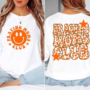 Può includere: T-shirt bianco con la scritta "PRAYING MOMS CLUB" in arancione, con una faccina sorridente sul davanti. Il retro della maglietta mostra lo stesso testo in un font retrò con accenti a stella. Il design incorpora una stampa animalier.