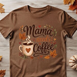 Può includere: Una t-shirt marrone a maniche corte con una stampa grafica che riporta il testo Mama needs Coffee. Il design include una tazza da caffè beige con un cuore, circondata da elementi autunnali come foglie d'acero arancioni e gialle e bacche rosse.