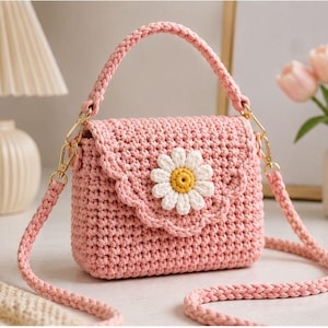 May include: A light pink crochet handbag featuring a scalloped flap closure and a central white daisy applique with a yellow center. The bag includes a braided top handle and a detachable matching braided shoulder strap with gold metal hardware.