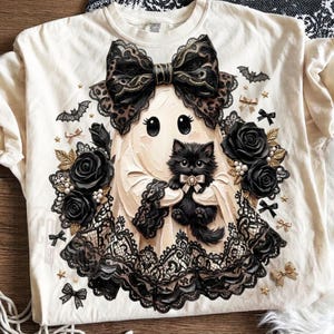 May include: A cream-colored t-shirt featuring a graphic of a ghost wearing a large black leopard-print bow, holding a fluffy black kitten. The design includes black lace trim, black roses, small white pearls, tiny gold stars, and small black bats.