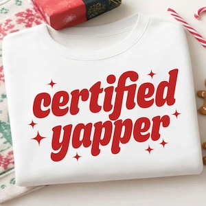 Könnte beinhalten: Weißes Sweatshirt mit rotem Schriftzug "certified yapper" und Sternen. Das Bild zeigt ein Geschenk, Zuckerstangen und Lebkuchenmänner, was auf ein weihnachtliches Thema hindeutet.