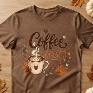 Könnte beinhalten: Ein braunes Kurzarm-T-Shirt mit einer Grafik einer Kaffeetasse mit Latte Art, umgeben von Herbstblättern und kleinen Herzen. Der Text "Coffee first" ist in einer dunkelbraunen Schreibschrift über der Tasse mit aufsteigendem Dampf gedruckt.